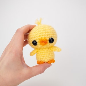 May include: A small yellow crocheted chick with black eyes and an orange beak. The chick is holding its wings out to the sides.