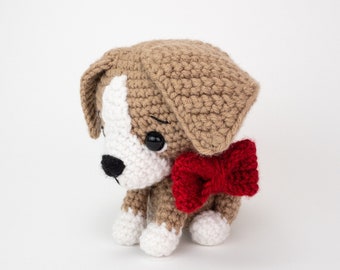 May include: A crocheted brown and white dog toy with a red bow. The dog has black eyes and a black nose.