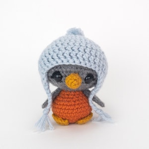 May include: A handmade crochet penguin doll with a gray head, orange body, and yellow feet. The penguin wears a light blue crocheted hat with ear flaps and a pom-pom. The doll has large black eyes and a yellow beak.