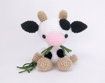 May include: A crocheted stuffed cow toy with black and white spots, a pink nose, and green yarn for a scarf. The toy is sitting on a white surface.