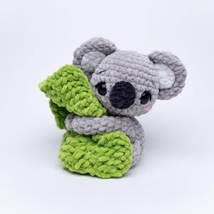 May include: A handmade grey crochet koala bear with a black nose, large black eyes, and pink cheeks. The koala is hugging a bright green, textured, crochet leaf. The ears have white inner details. The toy is on a white background.
