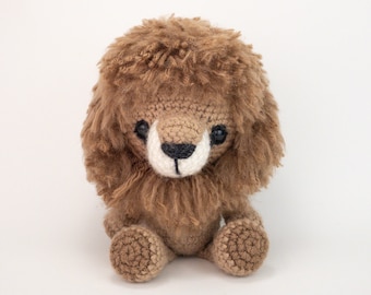 May include: A brown crocheted lion with a fluffy mane, black eyes, and a white nose. The lion is sitting with its legs crossed.