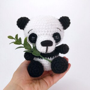 May include: A crocheted panda bear toy holding a green leaf. The panda is white with black ears, eyes, nose, and paws.