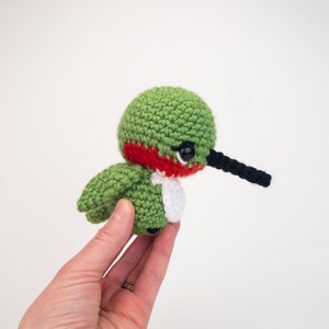May include: A green, red, and white crocheted hummingbird with black eyes and a long black beak.