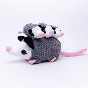 May include: A crocheted opossum with three baby opossums on its back. The opossum is grey with black ears, a white face, and a pink nose and tail. The baby opossums are white with black markings and pink noses.