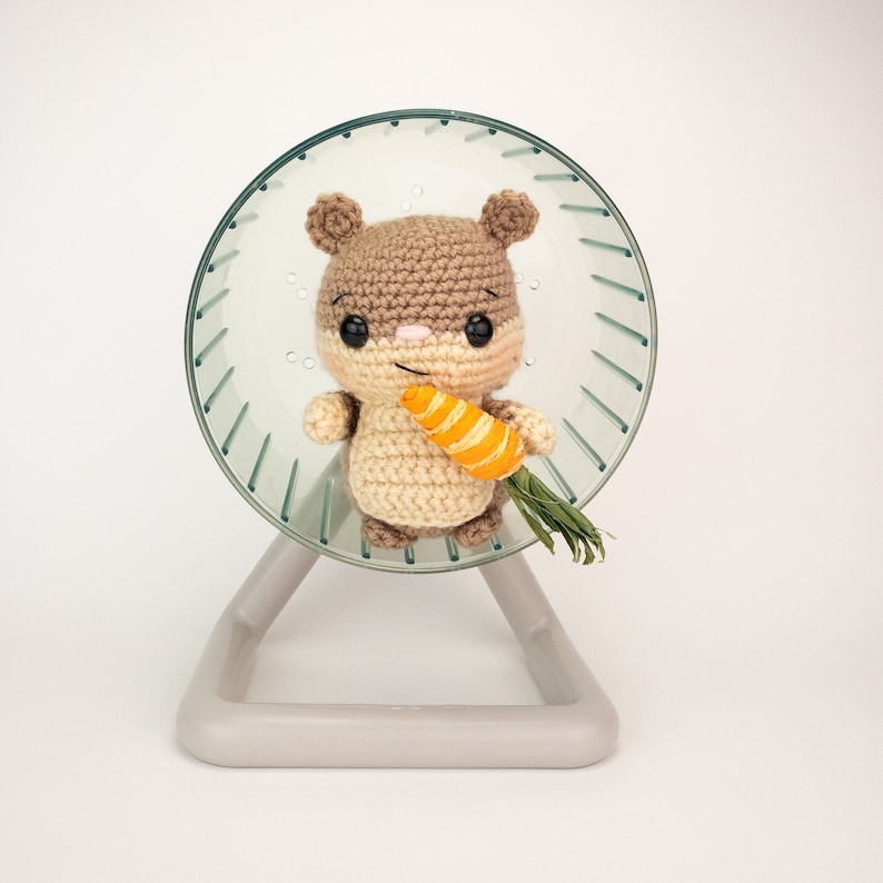 May include: A brown and white crocheted hamster holding a small orange carrot in a clear plastic hamster wheel.