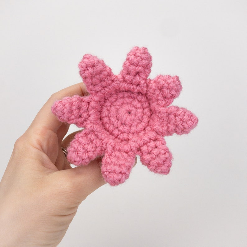 Puede incluir: Una flor de ganchillo rosa con ocho puntas, que se asemeja a una estrella de mar. La flor est&aacute; hecha a ganchillo en un patr&oacute;n circular con un c&iacute;rculo central.