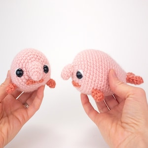 Peut inclure: Deux jouets en peluche en crochet rose et orange en forme de poisson-blob. Les jouets ont des yeux noirs et sont tenus par des mains.