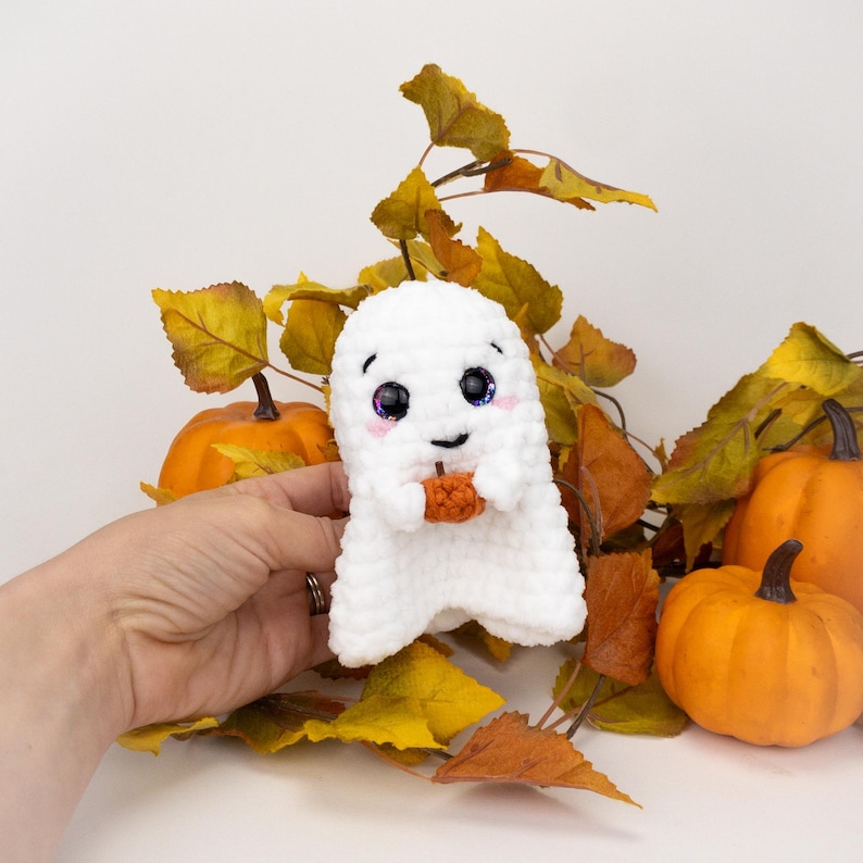 K&ouml;nnte beinhalten: Eine wei&szlig;e geh&auml;kelte Geisterpuppe mit gro&szlig;en, glitzernden Augen und einem kleinen orangefarbenen K&uuml;rbis. Der Geist h&auml;lt den K&uuml;rbis und ist von orangefarbenen K&uuml;rbissen und Herbstbl&auml;ttern umgeben. Eine niedliche Halloween-Dekoration.