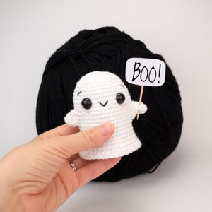 May include: A handmade white crochet ghost with black eyes and a smiling mouth, holding a sign that says "Boo!". The ghost is in front of a large ball of black yarn. A fun Halloween decoration.