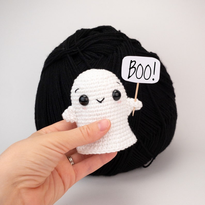 May include: A handmade white crochet ghost with black eyes and a smiling mouth, holding a sign that says "Boo!". The ghost is in front of a large ball of black yarn. A fun Halloween decoration.