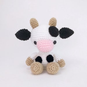 Puede incluir: Un juguete de peluche de vaca tejido a crochet con manchas blancas y negras, nariz rosa y pezu&ntilde;as marrones. La vaca est&aacute; sentada sobre un fondo blanco.