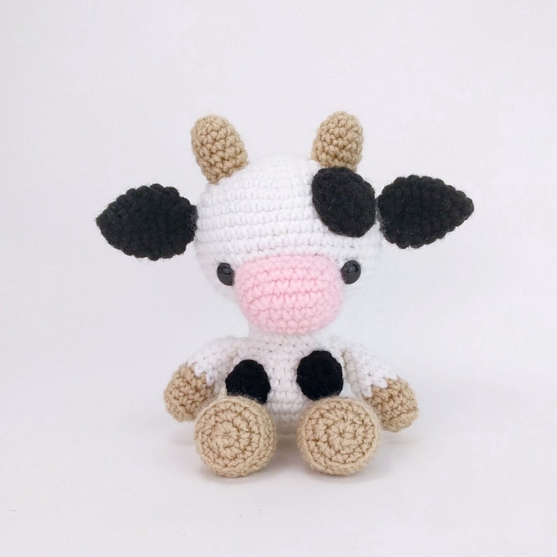 Puede incluir: Un juguete de peluche de vaca tejido a crochet con manchas blancas y negras, nariz rosa y pezu&ntilde;as marrones. La vaca est&aacute; sentada sobre un fondo blanco.
