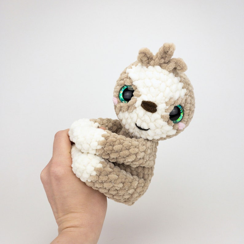 May include: A crocheted sloth plush toy with brown and white fur, green glitter eyes, and a pink nose. The sloth is holding its arms in front of it.