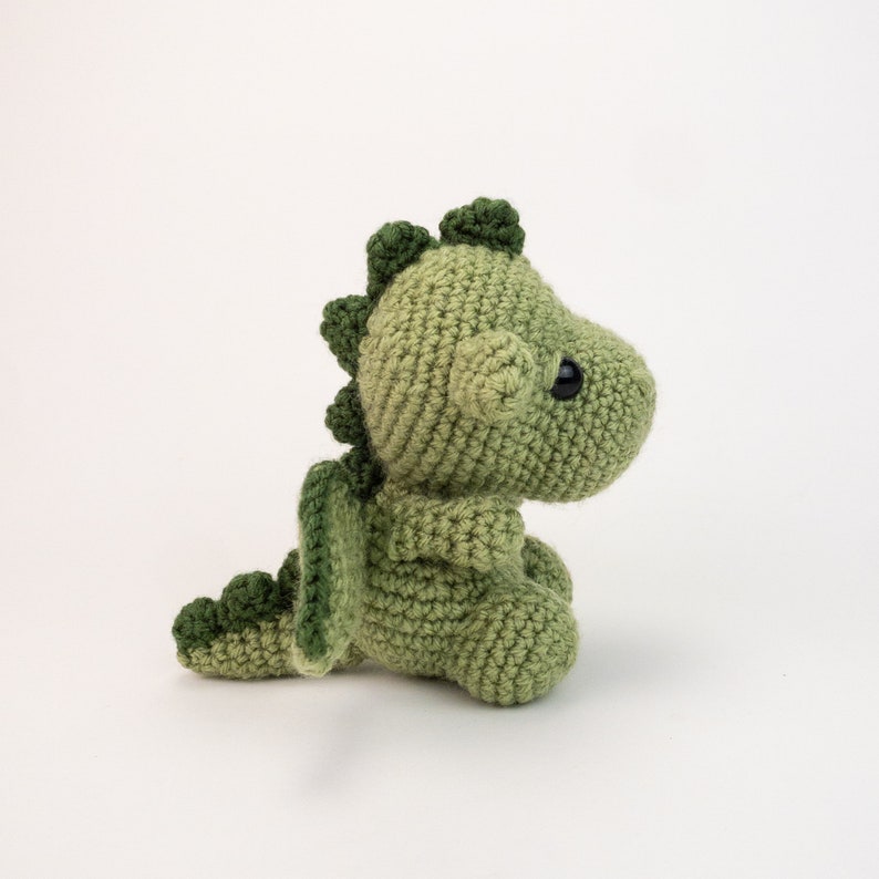 May include: A green crocheted dragon toy with a small tail and wings. The dragon has black eyes and a light green body.