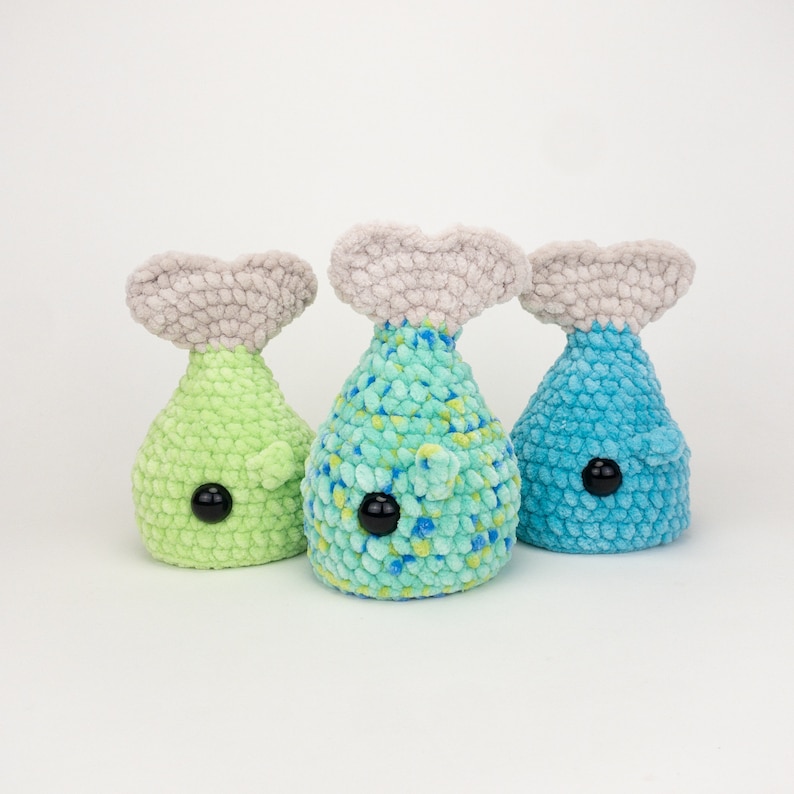 May include: Three crocheted whale plushies in different colors: green, blue, and teal. Each whale has a gray crocheted tail fin and black button eyes.