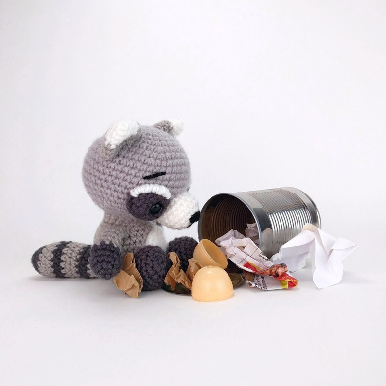 Puede incluir: Un juguete de peluche de mapache gris tejido a crochet se encuentra junto a una lata de metal volcada con papel arrugado y huevos de pl&aacute;stico en su interior.