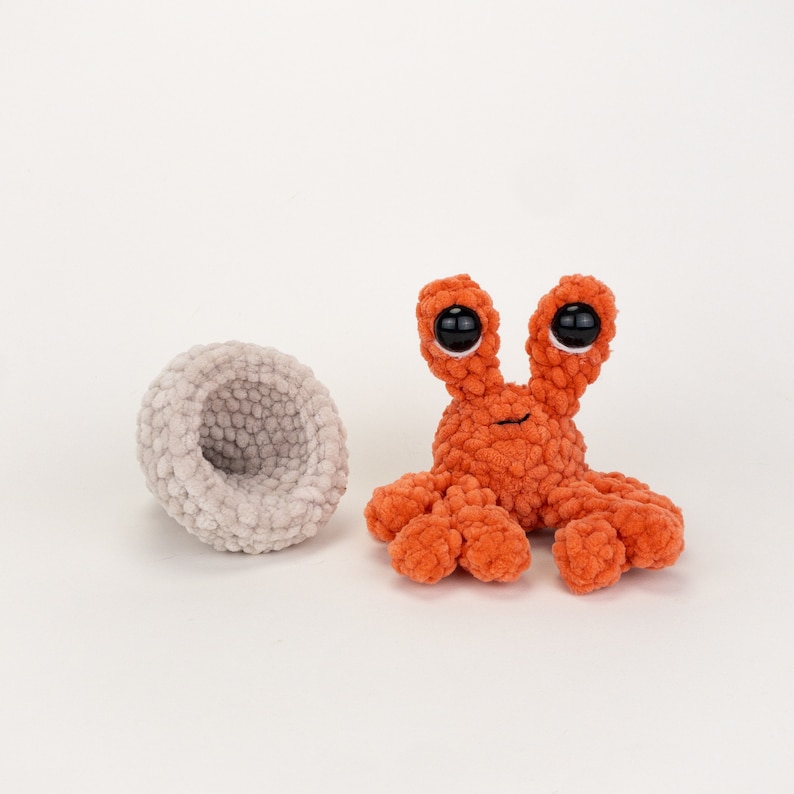 Może przedstawiać: Pomarańczowy krab na szydełku z dużymi czarnymi oczami i małym uśmiechem. Krab siedzi na białym tle obok szarej miski na szydełku.