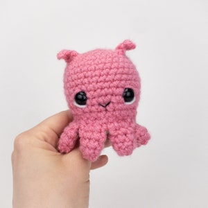 Puede incluir: Un pulpo de crochet rosa con ojos negros. El pulpo tiene un cuerpo pequeño y redondo y ocho tentáculos.