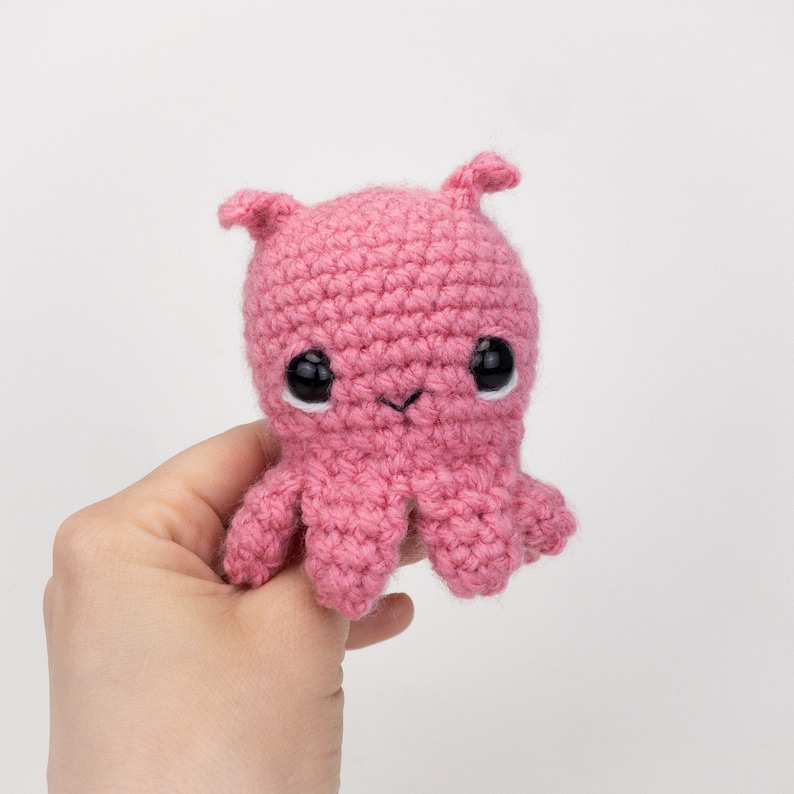 Puede incluir: Un pulpo de crochet rosa con ojos negros. El pulpo tiene un cuerpo peque&ntilde;o y redondo y ocho tent&aacute;culos.
