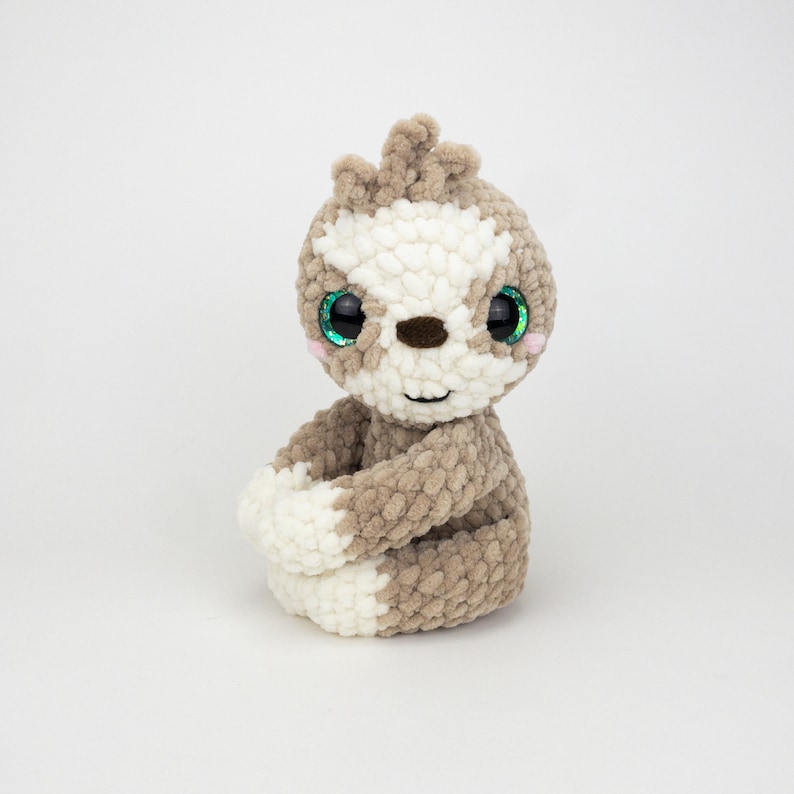May include: A crocheted sloth plush toy with brown and white fur, green eyes, and a pink nose. The sloth is sitting with its legs crossed.