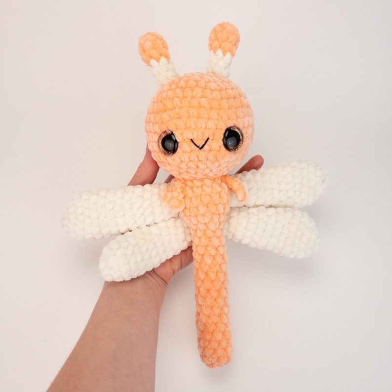 May include: A crocheted orange dragonfly plush toy with white wings. The dragonfly has large black eyes and a small smile.