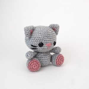 Puede incluir: Un juguete de crochet de gato gris con detalles rosas en las orejas y las patas. El gato tiene ojos negros y una nariz negra.