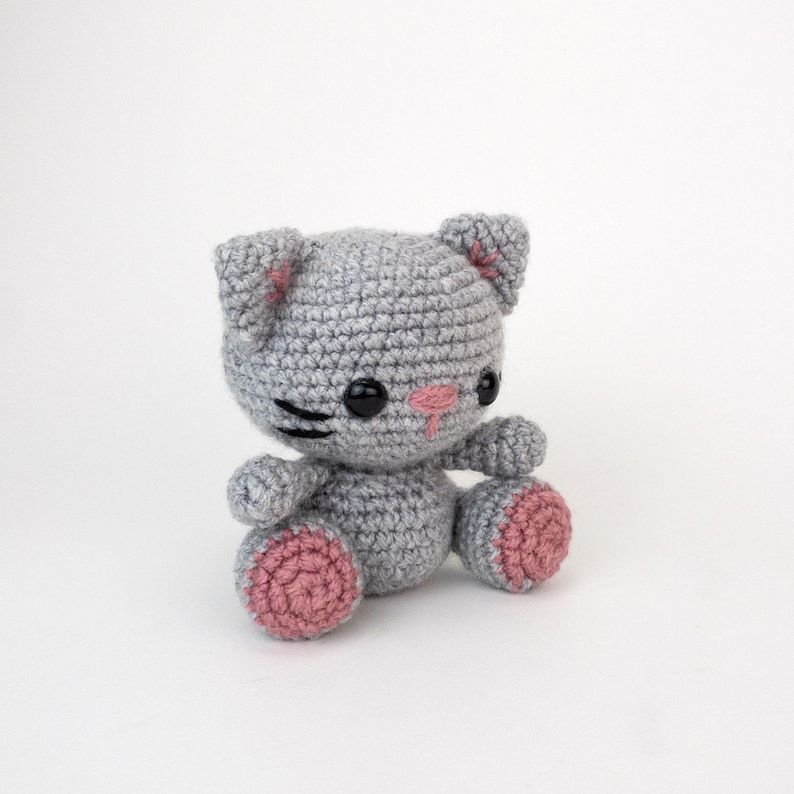 Puede incluir: Un juguete de crochet de gato gris con detalles rosas en las orejas y las patas. El gato tiene ojos negros y una nariz negra.