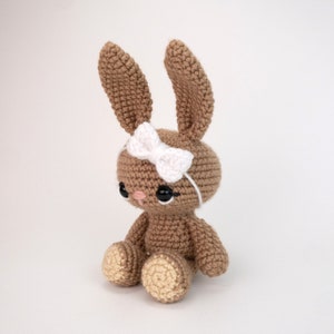 May include: A brown crocheted bunny rabbit with a white bow on its head. The bunny is sitting with its legs crossed and has black eyes.