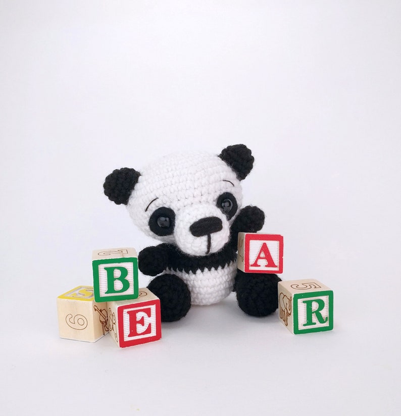 May include: A white and black crocheted panda bear toy sitting with wooden blocks that spell out "BEAR".