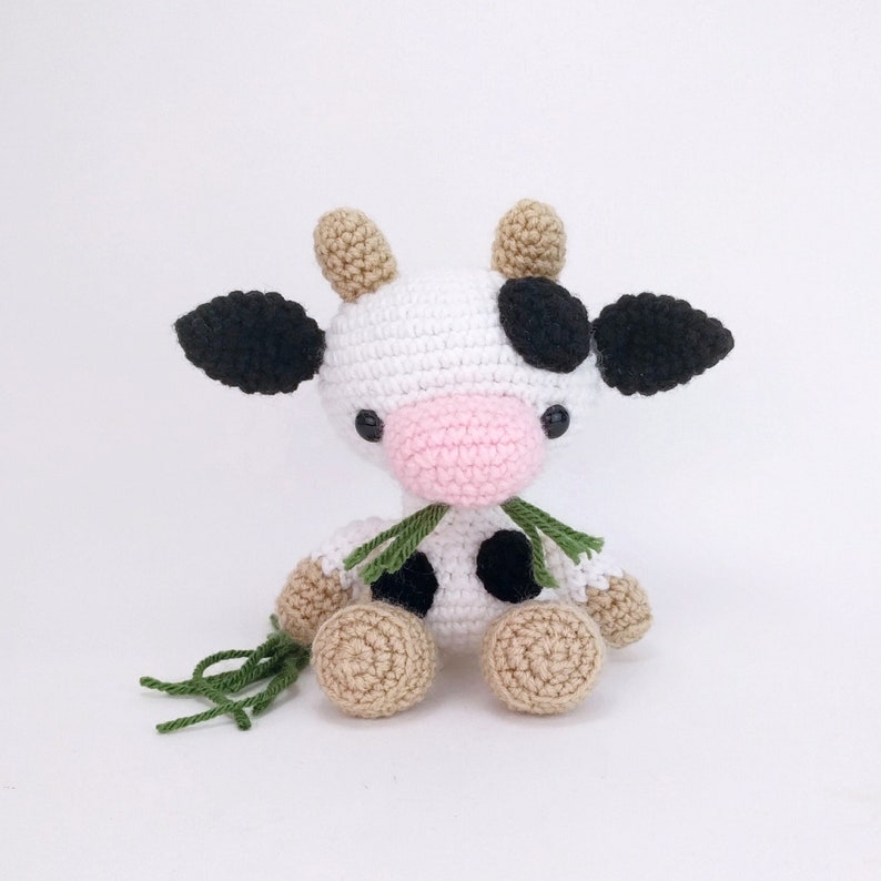 Puede incluir: Un juguete de peluche de vaca tejido a crochet con manchas blancas y negras, una nariz rosa y hilo verde para una bufanda. El juguete est&aacute; sentado sobre una superficie blanca.