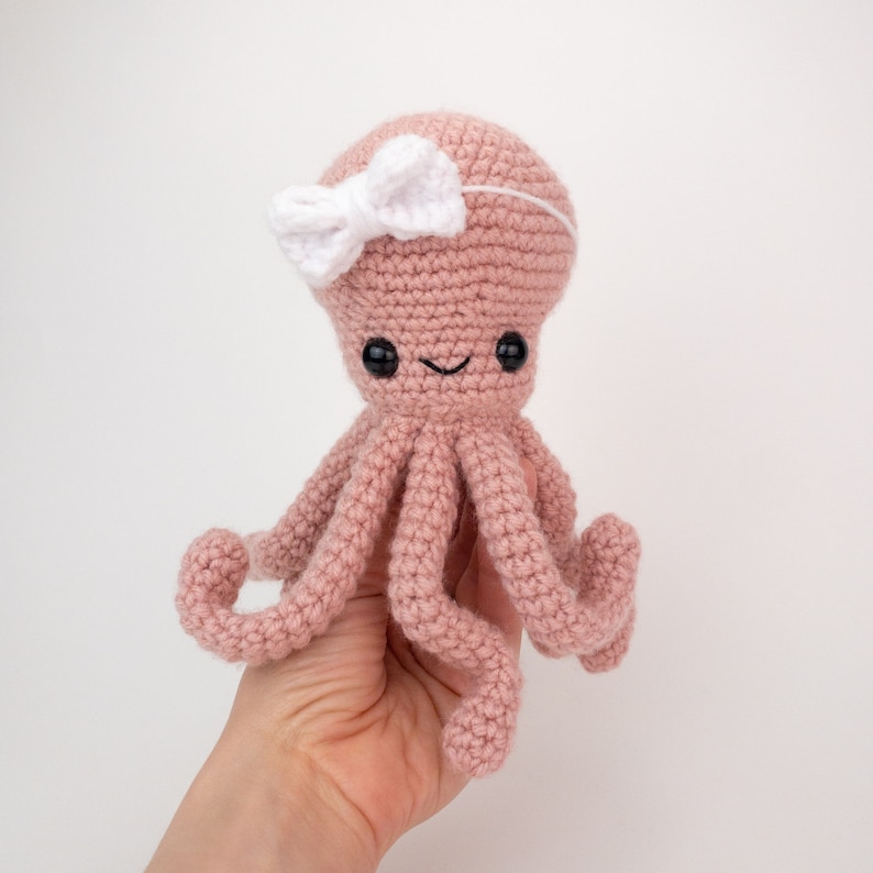 May include: A pink crocheted octopus with a white bow on its head. The octopus has black eyes and a smiling mouth.