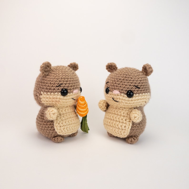 May include: Two brown crocheted stuffed hamsters, one holding a small orange carrot. The hamsters have black eyes and pink noses.