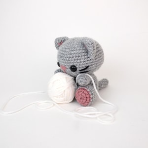 Puede incluir: Un juguete de crochet gris en forma de gato que sostiene una bola de hilo blanco. El gato tiene mejillas rosadas y ojos negros. El hilo se est&aacute; desenrollando.