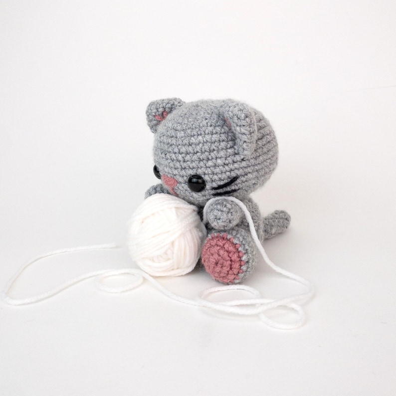 Puede incluir: Un juguete de crochet gris en forma de gato que sostiene una bola de hilo blanco. El gato tiene mejillas rosadas y ojos negros. El hilo se est&aacute; desenrollando.