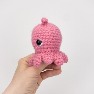 Puede incluir: Un pulpo de crochet rosa con ojos negros. El pulpo es peque&ntilde;o y tiene una expresi&oacute;n linda.