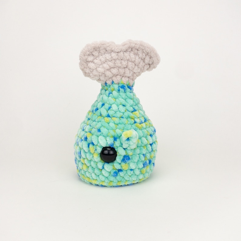 May include: A crocheted plush toy shaped like a whale tail. The whale is blue and green with a black eye. The tail fin is a light gray color.