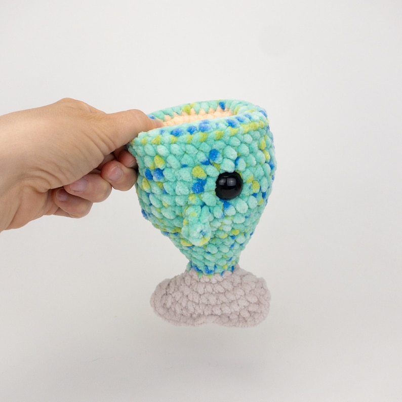 May include: A crocheted plush toy in the shape of a creature with a large head, a single black eye, and a small body. The head is a light blue color with yellow and green flecks. The body is a light gray color.