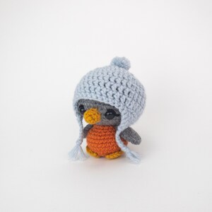 May include: A handmade crochet penguin doll with a light blue hat and orange body. The penguin has black button eyes and a yellow beak. The hat has ear flaps and a pom-pom on top. The penguin is approximately 4 inches tall.