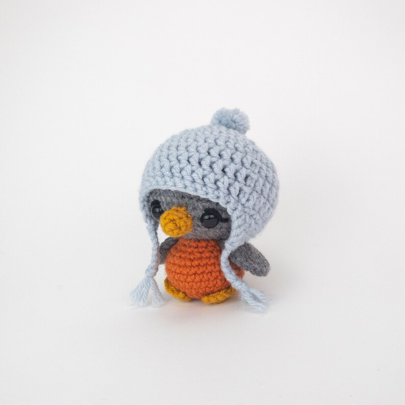 May include: A handmade crochet penguin doll with a light blue hat and orange body. The penguin has black button eyes and a yellow beak. The hat has ear flaps and a pom-pom on top. The penguin is approximately 4 inches tall.