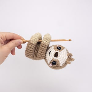 May include: A crocheted sloth toy hanging from a wooden stick. The sloth has brown and white fur, black eyes, and a pink nose.
