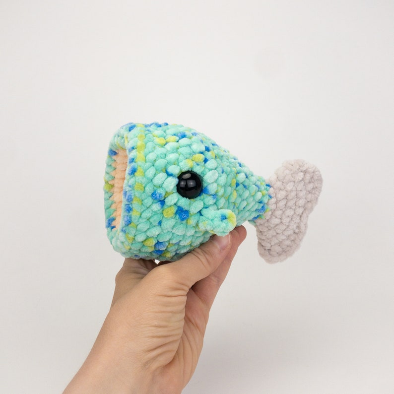 May include: A crocheted fish toy with a green and blue speckled body, a white tail fin, and a black eye.
