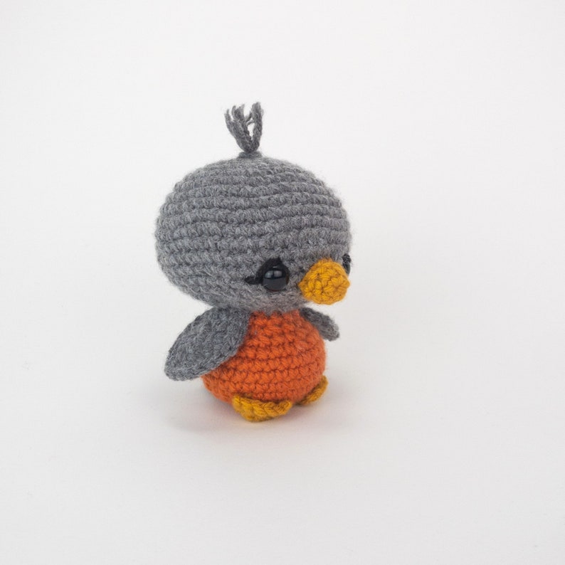 May include: A handmade crochet bird with a gray head, orange body, and yellow beak and feet. The small bird has black eyes and a small tuft of gray yarn on top of its head. The bird is a cute, whimsical design.