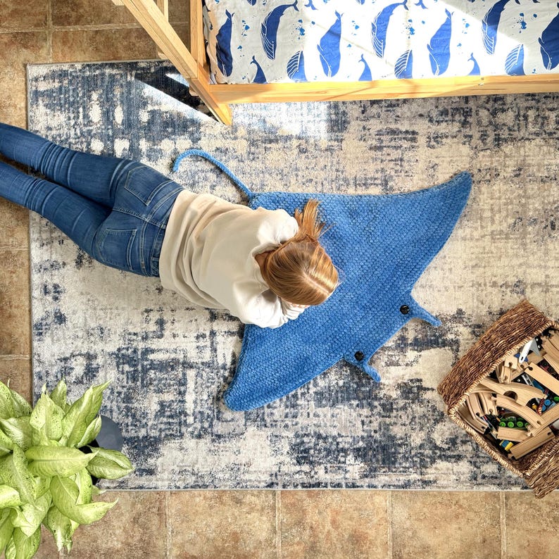 May include: A blue crocheted manta ray rug on a gray and white patterned rug. A person is lying on the rug with their head resting on the manta ray.