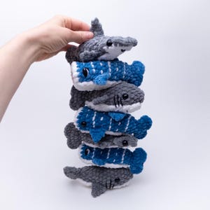 May include: A stack of six crocheted plush shark toys. The sharks are gray and blue with white accents. The sharks are stacked on top of each other, with the largest shark at the bottom and the smallest shark at the top.