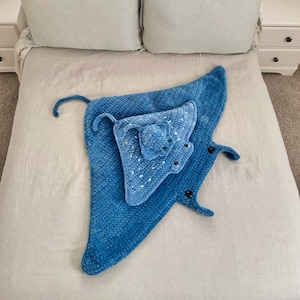 May include: A blue crocheted manta ray blanket with a smaller manta ray attached. The blanket is on a white bedspread.