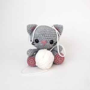 Puede incluir: Un juguete de crochet de gato gris con patas rosas sostiene una bola de lana blanca. El gato tiene ojos negros y una nariz rosa.