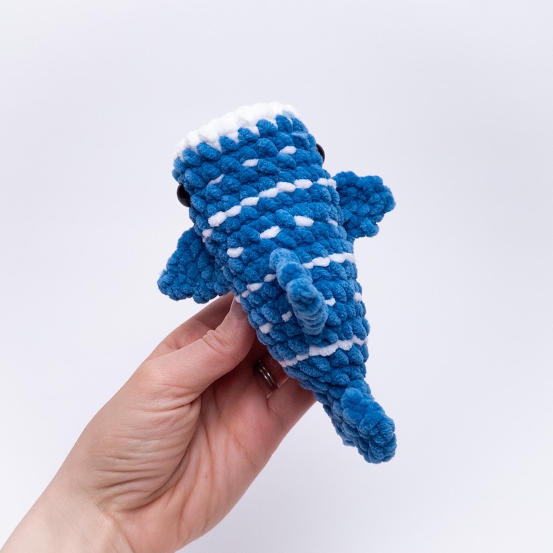 May include: A blue and white crocheted whale plush toy with black eyes. The whale is being held in a hand.