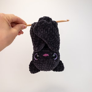 K&ouml;nnte beinhalten: Eine schwarze geh&auml;kelte Fledermaus, die kopf&uuml;ber h&auml;ngt, mit gro&szlig;en violetten Augen und einem rosa Mund.