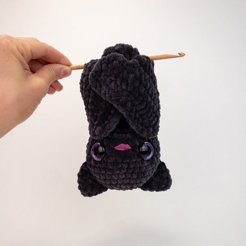 K&ouml;nnte beinhalten: Eine schwarze geh&auml;kelte Fledermaus, die kopf&uuml;ber h&auml;ngt, mit gro&szlig;en violetten Augen und einem rosa Mund.